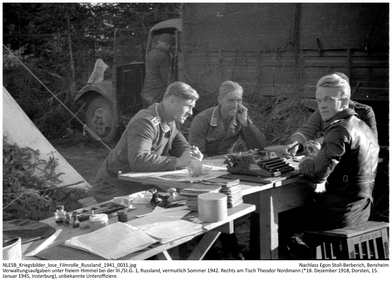Verwaltungsaufgaben - hier Abzeichnen der Leistungsbücher (Stapel auf dem vorderen Tisch) - unter freiem Himmel bei der III./St.G. 1, Russland, vermutlich Sommer 1942. Rechts am Tisch Theodor Nordmann (*18. Dezember 1918, Dorsten, 15. Januar 1945, Insterburg), unbekannte Unteroffiziere. NLESB_Kriegsbilder_lose_Filmrolle_Russland_1941_0031 Verwaltungsaufgaben - hier Abzeichnen der Leistungsbücher (Stapel auf dem vorderen Tisch) - unter freiem Himmel bei der III./St.G. 1, Russland, vermutlich Sommer 1942. Rechts am Tisch Theodor Nordmann (*18. Dezember 1918, Dorsten, 15. Januar 1945, Insterburg), unbekannte Unteroffiziere. NLESB_Kriegsbilder_lose_Filmrolle_Russland_1941_0031