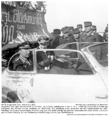 Hauptmann d.R. Egon Stoll-Berberich (17.06.1913 - 02.05.1973), Staffelkapitän in der III. SG 1,  erhält am 07.03.1944 in Orscha das Ritterkreuz des Eisernen Kreuzes (verliehen am 29.02.1944). Die Verleihung findet zusammen mit den Feierlichkeiten des 1000. Feindflugs des Gruppenkommandeurs Friedrich Lang statt. Die Staffel würdigt die geehrten Kameraden mit einem Banner und einem geschmückten Fahrzeug. Auf dem Beifahrersitz Friedrich Lang, dahinter Egon Stoll-Berberich. NLESB_Kriegsbilder_lose_Ritterkreuz_0011
